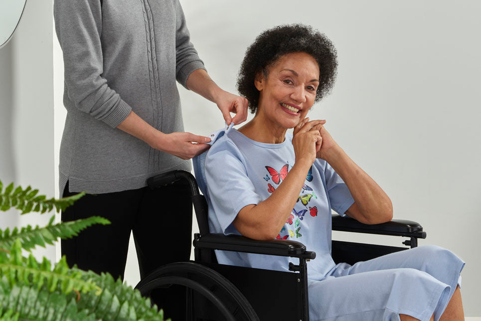 A woman in a wheelchair gets into her adaptive gown with the help of a caregiver.
