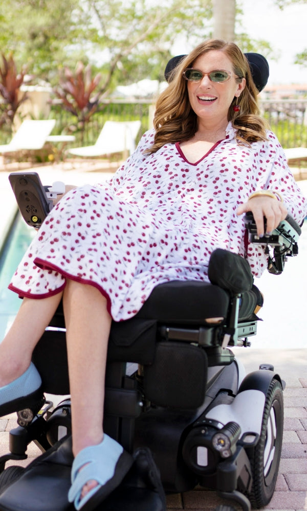 Woman in a wheelchair wearing a white cherry-print dress and light blue shoes outdoors.