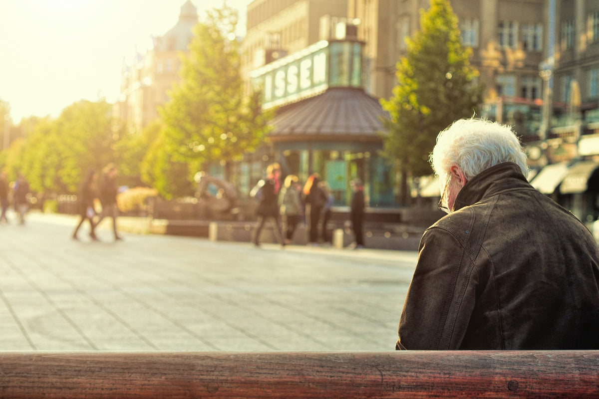 Older man sitting on a bench in a city. Looking downwards. People walking by at a distance.